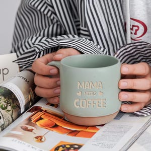 May include: A light green ceramic mug with the words "MAMA NEEDS COFFEE" etched into the side. The mug has a tan base and is held by a person wearing a striped shirt. The mug is sitting on top of a magazine.