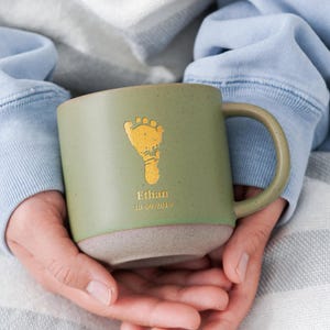 Pode incluir: Caneca de cerâmica verde com um design de pegada dourada e o nome "Ethan" com uma data. A caneca tem uma base cinza claro e uma alça curva. A caneca é segurada com as duas mãos.