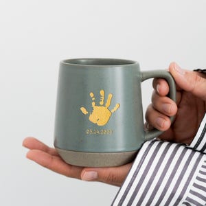 May include: A gray ceramic mug with a gold handprint and the date 03.14.2023. The mug has a gray handle and a tan base. The mug is held in a person's hand.