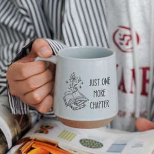 Peut inclure: Une tasse en céramique bleu clair avec une base marron, portant l'inscription "JUST ONE MORE CHAPTER" et une illustration florale et de livre. La tasse est tenue au-dessus d'un magazine ouvert.