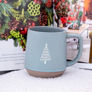 Puede incluir: Una taza de cerámica azul claro con un diseño de árbol de Navidad blanco. La taza tiene una base marrón y un asa curva. El fondo incluye una revista y decoraciones navideñas.