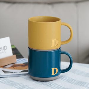 May include: Two ceramic mugs stacked on top of each other. The top mug is mustard yellow, and the bottom mug is teal. Each mug has a gold letter "D" on the front. The mugs are on a striped surface.