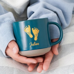 Puede incluir: Taza de cerámica color turquesa con base gris, con huellas doradas y el nombre "Julian". La taza es sostenida por una persona, con una sudadera azul y una manta a rayas en el fondo.