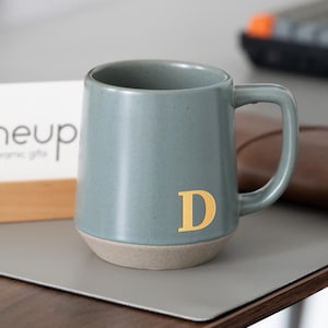 May include: A teal ceramic mug with a gold letter "D" on the front. The mug has a light tan base and a curved handle. The mug is on a gray surface.