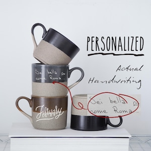 Puede incluir: Cuatro tazas de cerámica apiladas con mensajes escritos a mano. La taza superior es negra con un borde gris, la segunda taza es gris con un borde negro, la tercera taza es marrón con un borde gris y la taza inferior es negra con un borde gris. Las tazas están apiladas sobre un libro blanco. El texto en las tazas dice "Sei bella come Roma..." y "Febrero". El texto "PERSONALIZADO Escritura a mano real" está escrito en negro sobre fondo blanco.