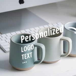 Peut inclure: Quatre mugs en céramique dans des tons de bleu et de vert avec une base marron clair. Les mugs ont une anse et le texte "Personnalisé LOGO TEXTE" est visible sur un mug. Les mugs sont sur une surface blanche.