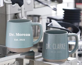 May include: Two blue-green ceramic mugs with a light brown base. One mug reads "Dr. Moreau Est. 2021" and the other reads "Dr. Clarke Ph.D. 2025" in white text. The mugs are on a stainless steel surface, with a coffee machine in the background.