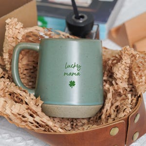 Puede incluir: Una taza de cerámica verde con las palabras "Lucky Mama" y un trébol verde. La taza está en una bandeja de cuero marrón llena de papel marrón. Una caja marrón y una revista están en el fondo.