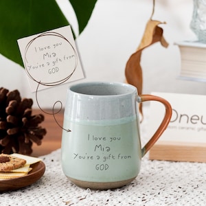 May include: A ceramic mug with a light blue and gray design, featuring a brown handle. The mug has the handwritten text "I love you Mia, You're a gift from GOD". A small card with the same text is also present.