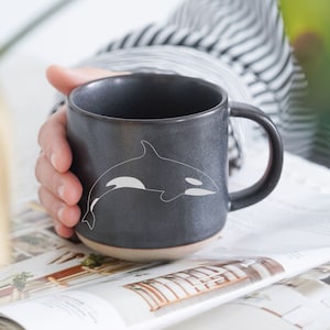 Op de afbeelding: Een zwarte keramische mok met een wit orka-ontwerp. De mok heeft een afgeronde handgreep en een licht uitlopende rand.