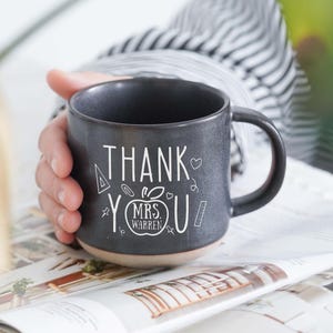 Puede incluir: Una taza de cerámica gris oscuro con base beige, sostenida por una persona. La taza presenta texto blanco e ilustraciones que dicen "THANK YOU MRS. WARREN". El diseño incluye un corazón, estrellas e iconos escolares. La taza está sobre una revista.