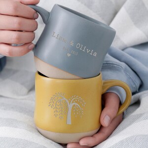 May include: Two ceramic mugs, one stacked on top of the other. The top mug is a dusty blue with the engraved text "Liam & Olivia Est. 2015". The bottom mug is yellow with a tree design.