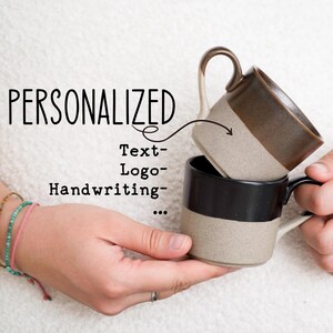 Könnte beinhalten: Zwei Keramikbecher, einer auf dem anderen gestapelt, werden in Händen gehalten. Der obere Becher ist braun und beige, der untere ist schwarz und beige. Das Wort "PERSONALIZED" steht über den Bechern, darunter "Text-Logo-Handschrift".