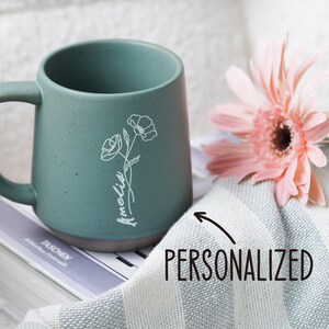 May include: A personalized green ceramic mug with a white floral design and the name "Amelia" written on it. The mug is sitting on a book and a white and gray striped towel. A pink flower is also in the image.
