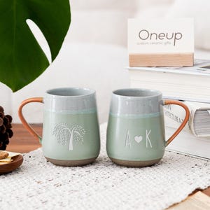 May include: Two ceramic mugs with light green and gray glaze, featuring brown handles. One mug has a tree design, the other has "A♥K". A wooden sign reads "Oneup custom ceramic gifts". A small wooden plate holds crackers.