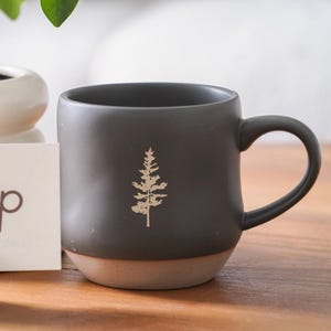 Puede incluir: Una taza de cerámica gris oscuro con base y asa marrón claro. La taza presenta un diseño de árbol dorado en la parte delantera. La taza está sobre una superficie de madera junto a una caja blanca con el texto "Oneup custom ceramic gifts".