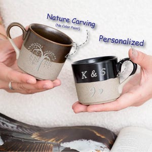 May include: Two ceramic mugs with a two-tone design. One mug is brown with a tree carving, the other is black with the initials "K & S" and a heart. Both mugs have a tan base and black handles.