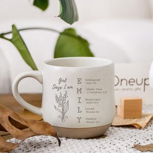 Puede incluir: Una taza de cerámica blanca moteada con base marrón, que presenta un dibujo lineal negro de flores de lirio de los valles y el texto God Says I am EMILY con los versículos bíblicos correspondientes para cada letra: Embraced, Made New, Important, Loved, Yielded.