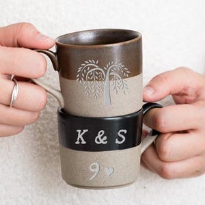 May include: Two stacked ceramic mugs. The top mug is brown and beige with a tree design. The bottom mug is black and beige with the letters "K & S" and the number "9" with a heart symbol. The mugs are held by a person.