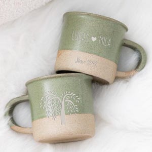 Op de afbeelding: Twee groene keramische mokken met bruinbeige bodems en handvatten zijn op een witte, zachte ondergrond gestapeld. Op één mok staan de namen "LUCAS ️ MILA" en "Since 2016" gegraveerd. De andere mok heeft een boomontwerp.