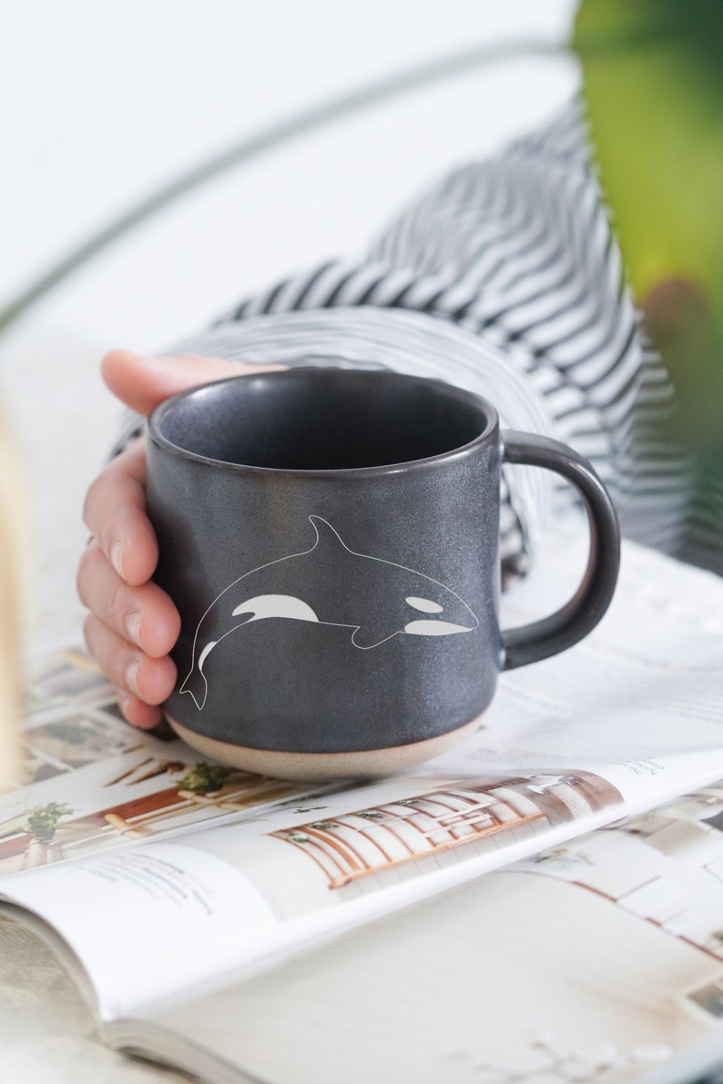 Pu&ograve; includere: Una tazza in ceramica nera con un design di orca bianca. La tazza ha un manico arrotondato e un bordo leggermente svasato.