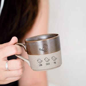 May include: A two-tone ceramic mug with a brown top and a beige bottom. The mug features a bird illustration and the names "Luna", "Chloe", and "Mia" printed below three additional bird illustrations. The mug has a curved handle.