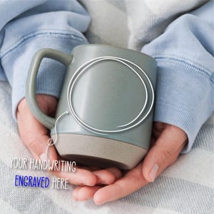 May include: A speckled, light blue ceramic mug with a tan base and a large handle. The mug is held in two hands. Text on the image reads "YOUR HANDWRITING ENGRAVED HERE". A white circle is drawn on the mug.