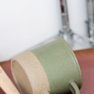 May include: A ceramic mug with a two-tone design, featuring a speckled sage green upper half and a tan lower half. The mug has a curved handle and rests on a wooden spoon. The mug is approximately 4 inches tall.