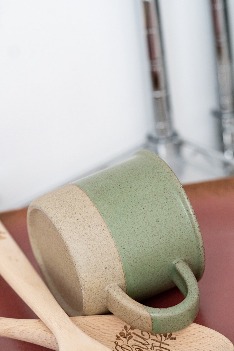 May include: A ceramic mug with a two-tone design, featuring a speckled sage green upper half and a tan lower half. The mug has a curved handle and rests on a wooden spoon. The mug is approximately 4 inches tall.