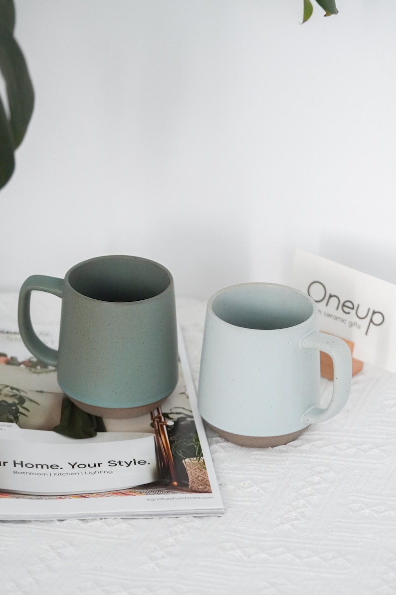May include: Two ceramic mugs in the image. One is a muted teal color, the other is a light blue. Both have a brown base and a handle. The mugs are on a magazine with the text "Your Home. Your Style."