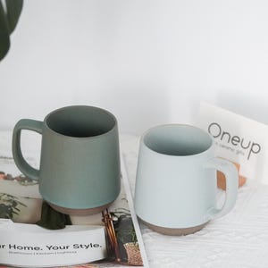 May include: A set of three ceramic mugs with a speckled, matte finish. The mugs are in shades of cream, light gray, and dark gray. Each mug has a simple, minimalist design and a rounded handle.