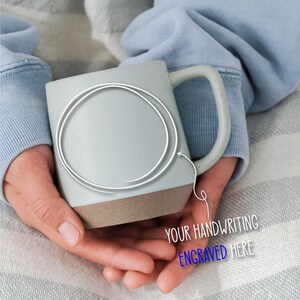 May include: A light gray ceramic mug with a brown base is held in both hands. The mug has a large handle and a white circle with an arrow pointing to the mug. Text on the image reads "YOUR HANDWRITING ENGRAVED HERE".