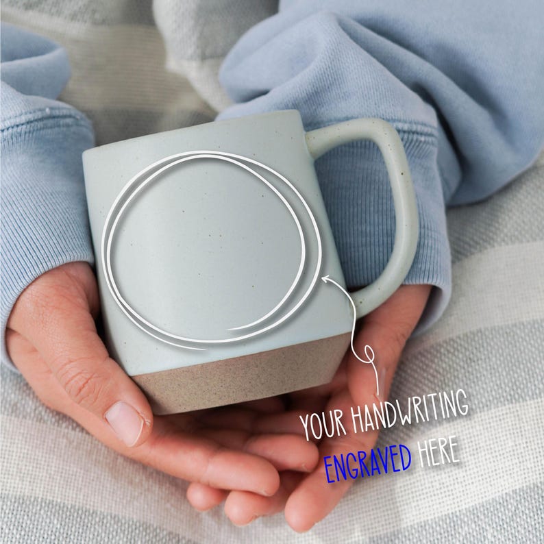May include: A light gray ceramic mug with a brown base is held in both hands. The mug has a large handle and a white circle with an arrow pointing to the mug. Text on the image reads "YOUR HANDWRITING ENGRAVED HERE".