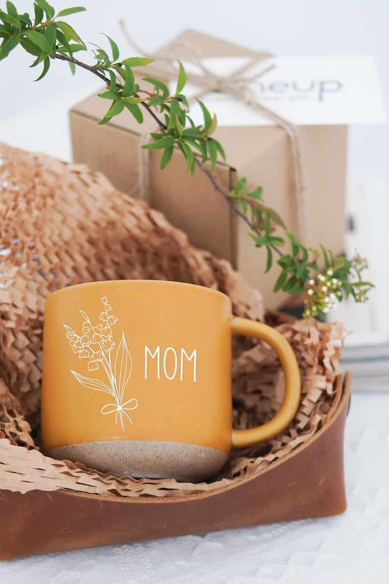 May include: Mustard yellow ceramic mug with the word "MOM" and a floral design in white. The mug sits in a brown leather container with brown paper filler, with a gift box in the background. Green sprigs add a natural touch.