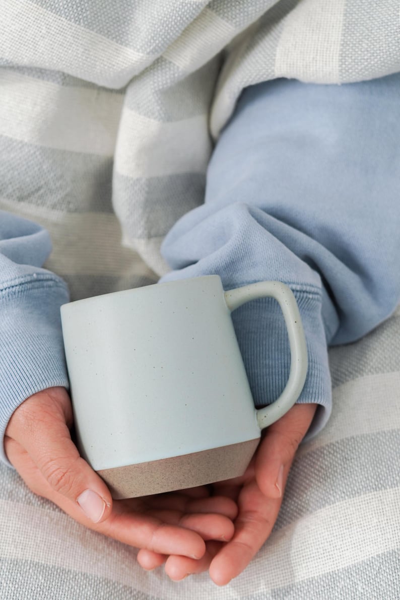 May include: A light blue ceramic mug with a brown base is held in both hands. The mug has a simple design and a curved handle. The person holding the mug is wearing a light blue sweatshirt.