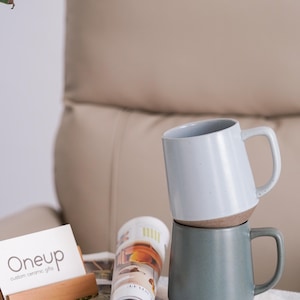 May include: Two ceramic mugs, one light blue and one teal, stacked on top of each other. The mugs have a two-tone design with a brown base. A magazine and a wooden business card holder with the text "Oneup custom ceramic gifts" are in the foreground.
