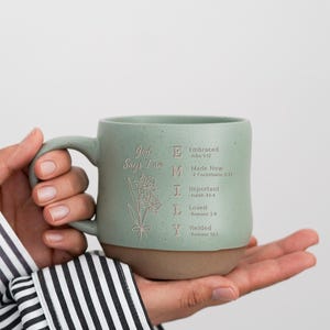 Puede incluir: Una taza de cerámica verde salvia con acabado moteado y base marrón. La taza presenta un dibujo floral de líneas y el texto: God Says I am E Embraced-John 1:12, M Made New-2 Corinthians 5:17, I Important-Isaiah 43:4, L Loved-Romans 5:8, Y Yielded-Romans 12:1.