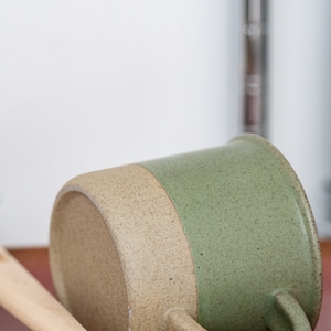 May include: A handmade ceramic mug with a speckled, two-tone design. The bottom half is a light tan color, while the top half is a muted green. The mug has a curved handle and rests on wooden utensils.
