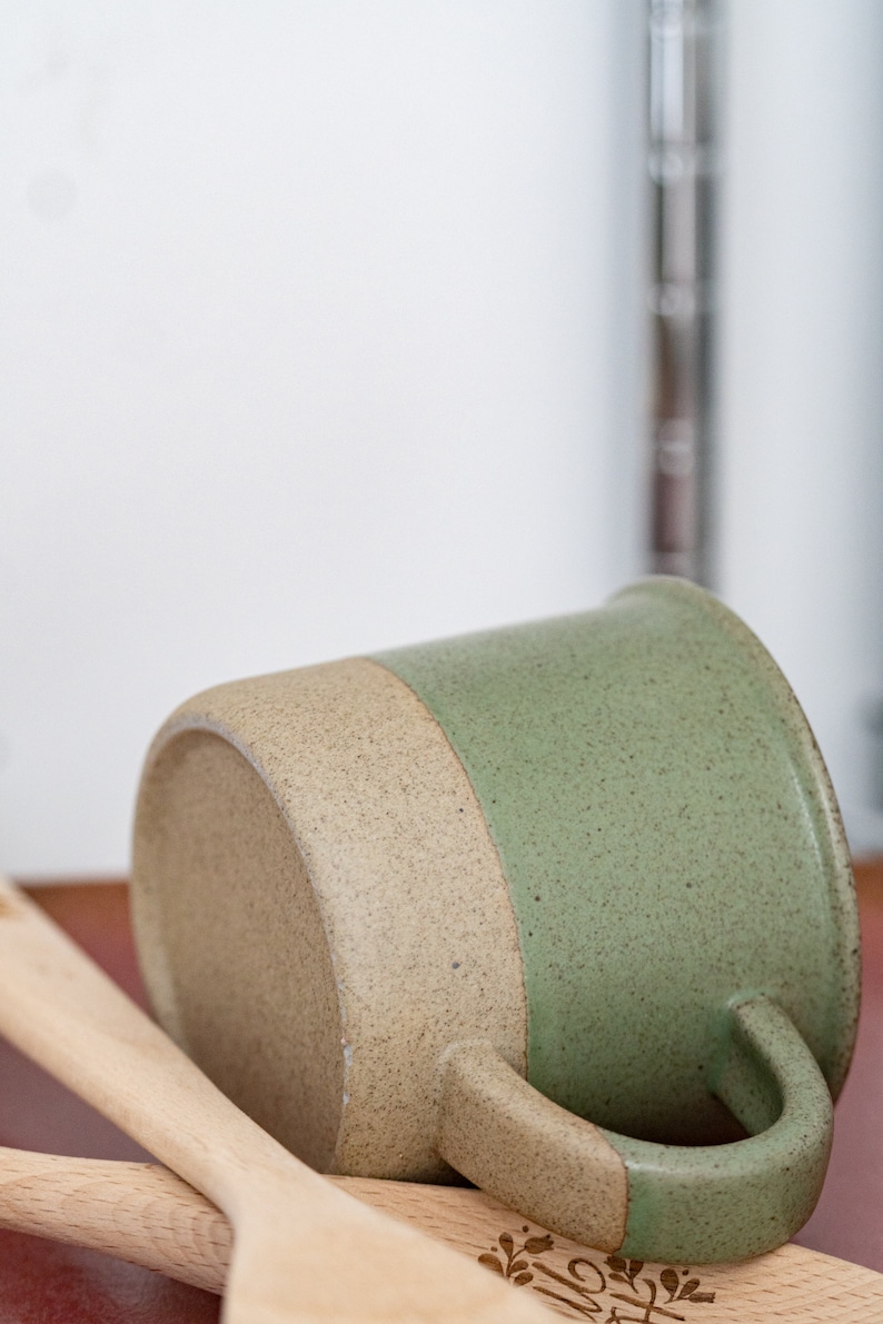 May include: A handmade ceramic mug with a speckled, two-tone design. The bottom half is a light tan color, while the top half is a muted green. The mug has a curved handle and rests on wooden utensils.
