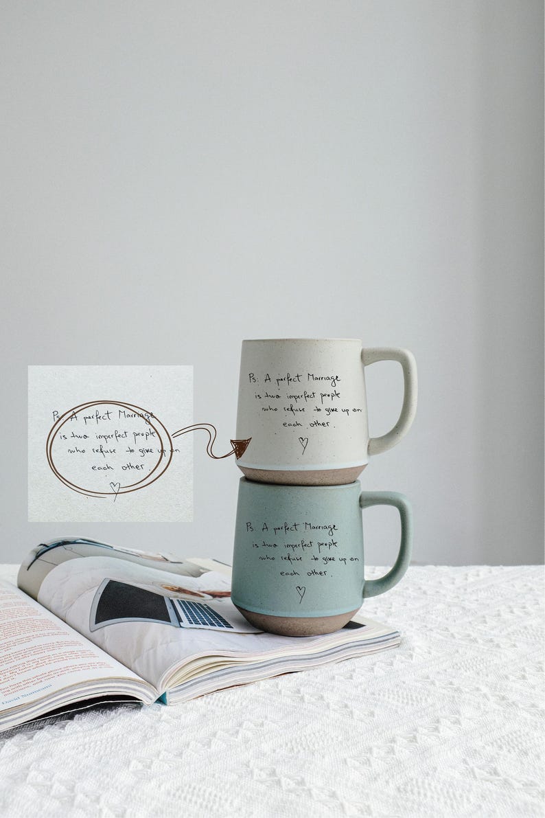 May include: Two ceramic mugs, one cream and one teal, stacked on a magazine. Each mug has a brown base and a handle. The text "A perfect Marriage is two imperfect people who refuse to give up on each other" is written on each mug.