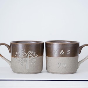 May include: Two ceramic mugs in brown and grey, one with a tree design, the other with the initials "K & S" and a heart. Both mugs have a curved handle. Ideal for a couple.