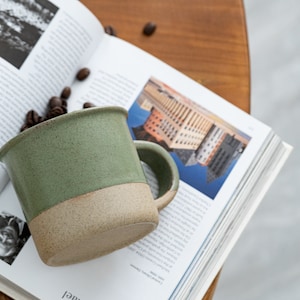 May include: A handmade ceramic mug with a green upper half and a tan lower half, filled with coffee beans. The mug sits on an open book with black and white images and text. The mug has a rounded handle.