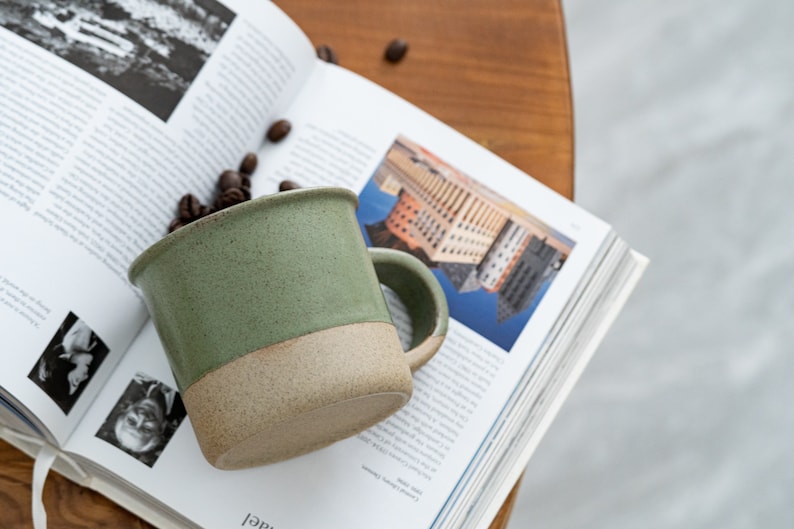 May include: A handmade ceramic mug with a green upper half and a tan lower half, filled with coffee beans. The mug sits on an open book with black and white images and text. The mug has a rounded handle.