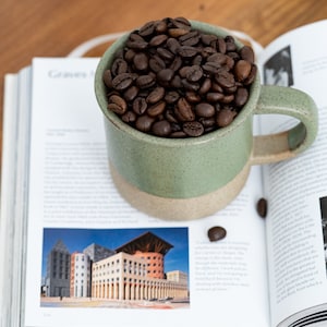 May include: A green and beige ceramic mug filled with roasted coffee beans, resting on an open book. The book features architectural images and text. The mug is on a wooden surface.