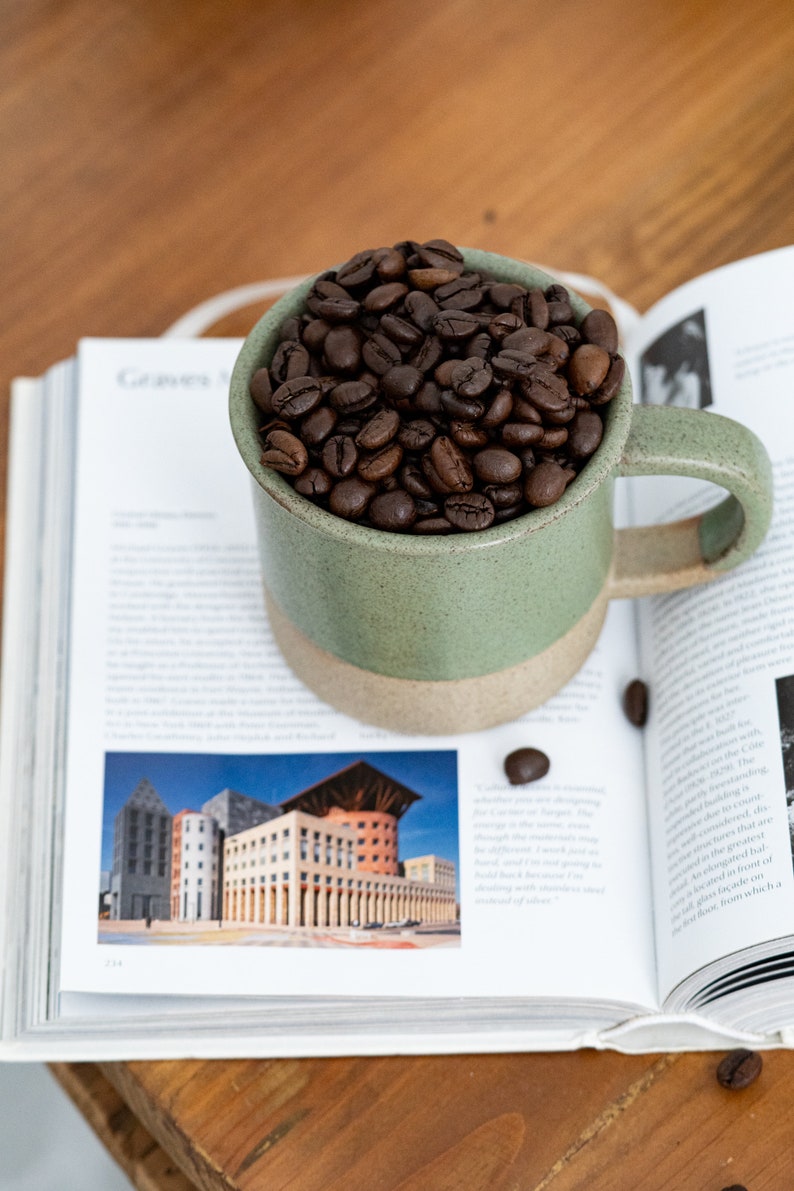 May include: A green and beige ceramic mug filled with roasted coffee beans, resting on an open book. The book features architectural images and text. The mug is on a wooden surface.