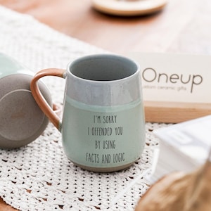 May include: A ceramic mug with a light green base and gray top, featuring a brown handle. The mug has the text "I'M SORRY I OFFENDED YOU BY USING FACTS AND LOGIC".