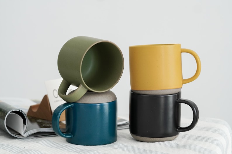May include: Four ceramic mugs in shades of green, teal, yellow, and black are stacked. Each mug has a contrasting grey base and a curved handle. The mugs are on a white surface with a magazine and a wooden object in the background.
