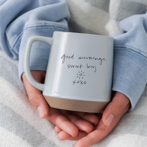 以下が含まれることがあります： 「good morning, sweet boy」の文字と太陽、「xoxo」が手書きで書かれた、茶色の底を持つ白いセラミックマグカップ。大きなハンドルが付いており、両手で持たれています。