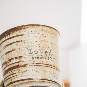 Peut inclure: Un mug cylindrique en céramique avec un émail crème moucheté et des motifs de coups de pinceau bruns horizontaux. Le texte "Loved. -Romans 5:8" est imprimé dans une police sombre de style machine à écrire sur le devant.