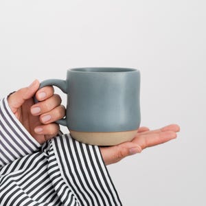 May include: A person's hand holding a gray ceramic mug with a light brown rim. The mug is slightly wider at the top than the bottom.